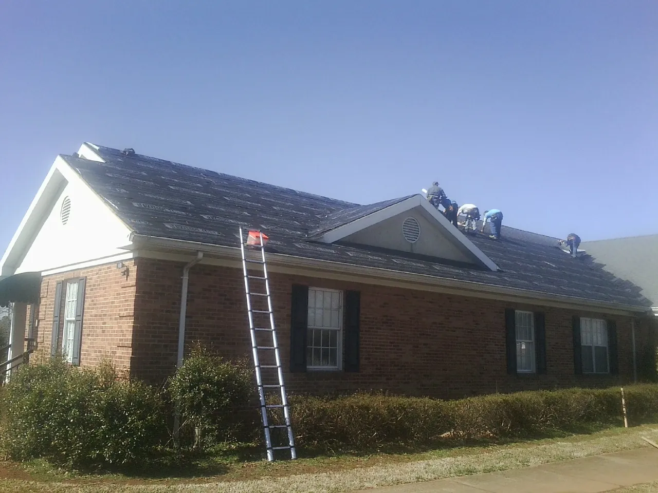 Crew tearing shingles off a brick ranch roof with a ladder set — Emergency Roof Repair work by our crew in Cedar Knolls
