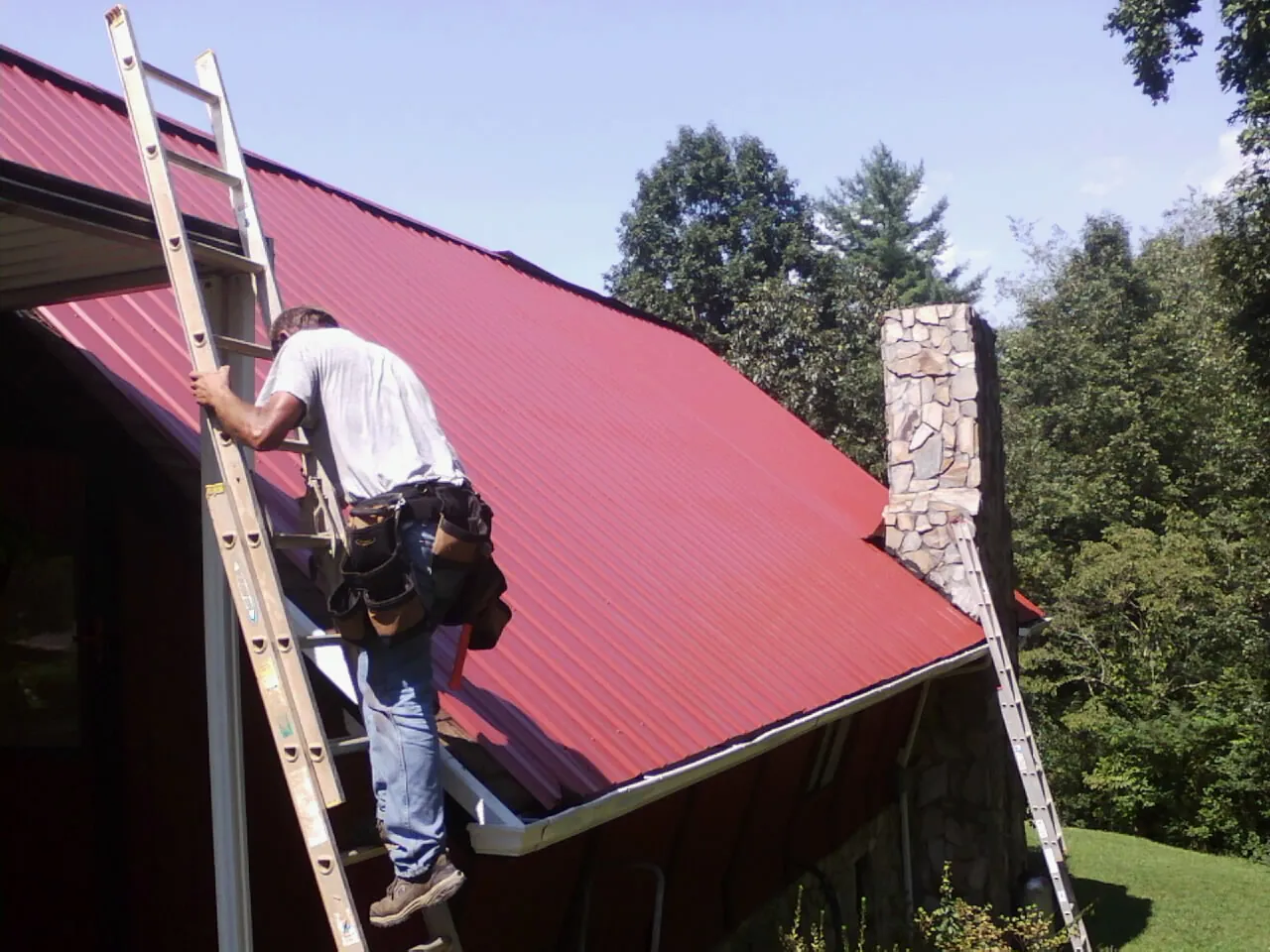 Roofer on a ladder beside a red metal roof and stone chimney — Soffit & Fascia Repair by Roofing Brothers in Cedar Knolls